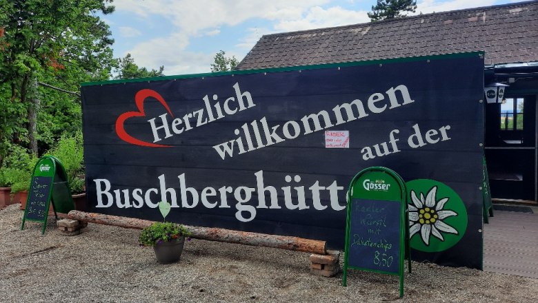 Herzlich Willkommen, © Buschberghütte Willkommensschild der Buschberghütte mit grüner Umgebung und blauen Himmel.