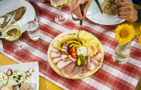 Heurigenjause, © POV / Robert Herbst Ein gedeckter Tisch mit einer Platte voller Aufschnitt, Käse und Gemüse, dazu Weißwein und Brot auf einer rot-weiß karierten Tischdecke.