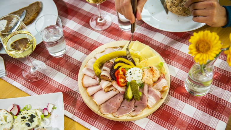 Heurigenjause, © POV / Robert Herbst Ein gedeckter Tisch mit einer Platte voller Aufschnitt, Käse und Gemüse, dazu Weißwein und Brot auf einer rot-weiß karierten Tischdecke.