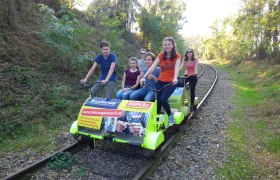Weinvierteldraisine, © Weinvierteldraisine Gruppe von Menschen fährt auf einer Draisine auf Bahngleisen durch eine bewaldete Landschaft.