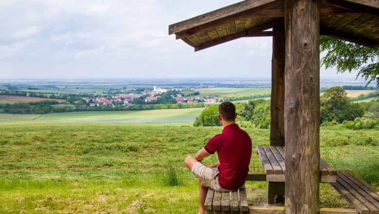 Panoramic view, © Fotostudio Semrad A man sits under a wooden shelter and looks out over a wide landscape with fields and a village in the distance.