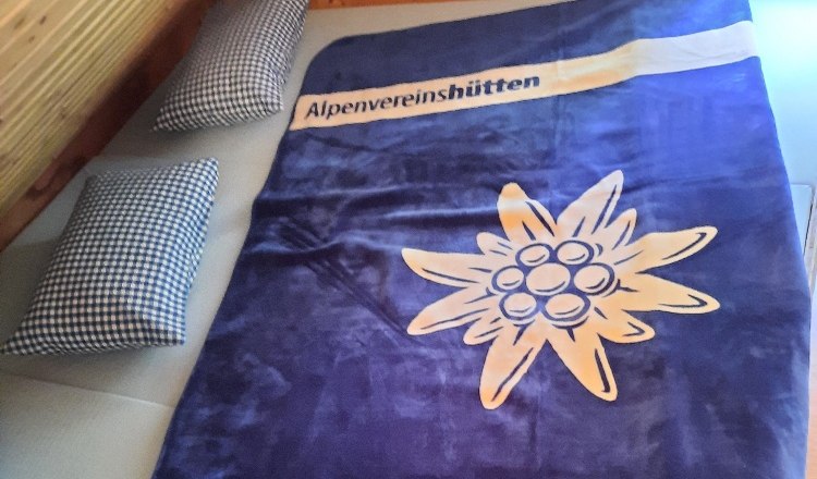 Matratzenlager, © Buschberghütte Ein Matratzenlager mit einer blauen Decke, die ein Edelweiß und den Schriftzug 'Alpenvereinshütten' zeigt, sowie zwei karierte Kissen.