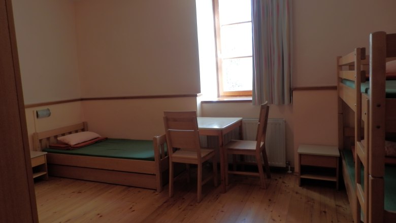 Gästezimmer, © Geistliches Jugendzentrum Oberleis Ein einfach eingerichtetes Gästezimmer mit Holzboden, Einzelbett, Etagenbett, Tisch und Stühlen, sowie einem Fenster mit Vorhang.