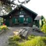 Buschberghütte, © Buschberghütte Eine rustikale Holzhütte mit grünen Fensterläden und Blumenkästen, umgeben von Bäumen und Gartenmöbeln.