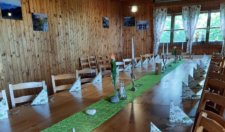 Innenbereich, © Buschberghütte Ein rustikaler Raum mit Holzwänden und -decke. Ein langer Tisch ist mit grüner Tischdecke, Kerzen und gefalteten Servietten gedeckt.