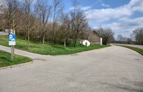 Wohnmobilstellplatz Wenzersdorf, © Gemeinde Gnadendorf Parkplatz mit Wohnmobilstellplatzschild, Bäume und altes Gebäude im Hintergrund.