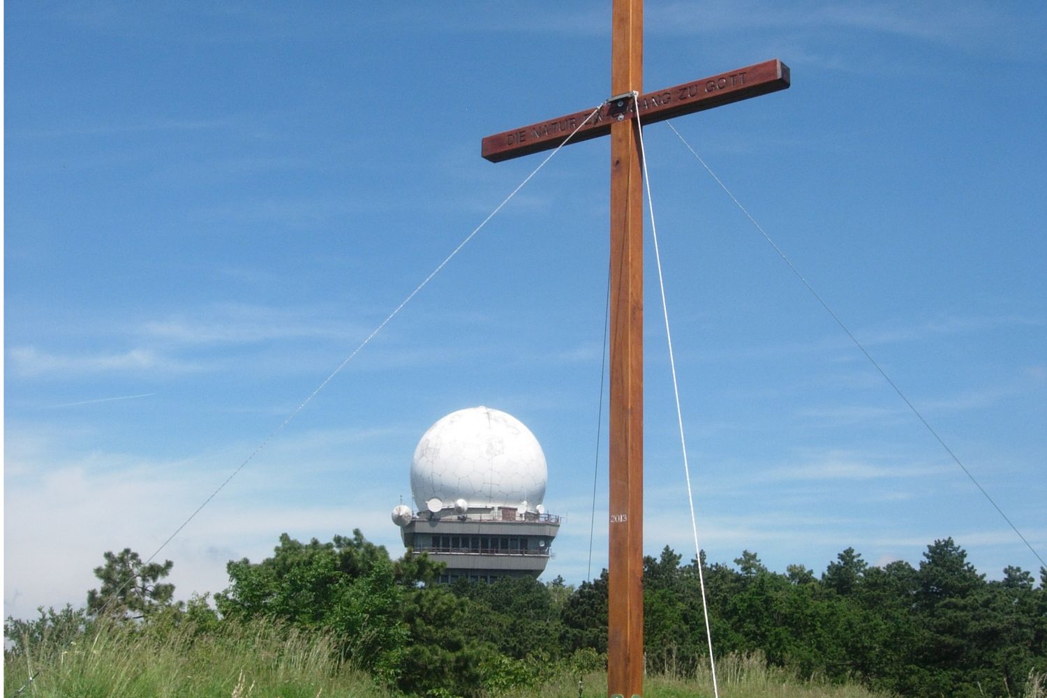 Ein Holzkreuz vor einer Radarkuppel auf dem Buschberg, umgeben von Bäumen und blauem Himmel.