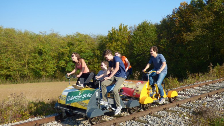 Gruppe von Menschen f&auml;hrt auf einer Draisine auf Bahngleisen durch eine l&auml;ndliche Landschaft.