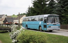 Ein blauer Bus fährt durch ein Dorf mit Blumen und Bäumen im Hintergrund.