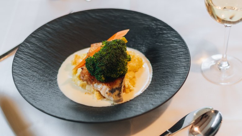 Ein elegant angerichtetes Gericht auf einem schwarzen Teller mit Gem&uuml;se und Sauce, daneben ein Glas Wei&szlig;wein auf einem gedeckten Tisch.