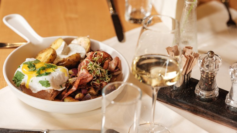 Ein Teller mit einem Gericht aus Fleisch, Kartoffeln und Spiegelei, serviert mit einem Glas Wei&szlig;wein auf einem gedeckten Tisch.