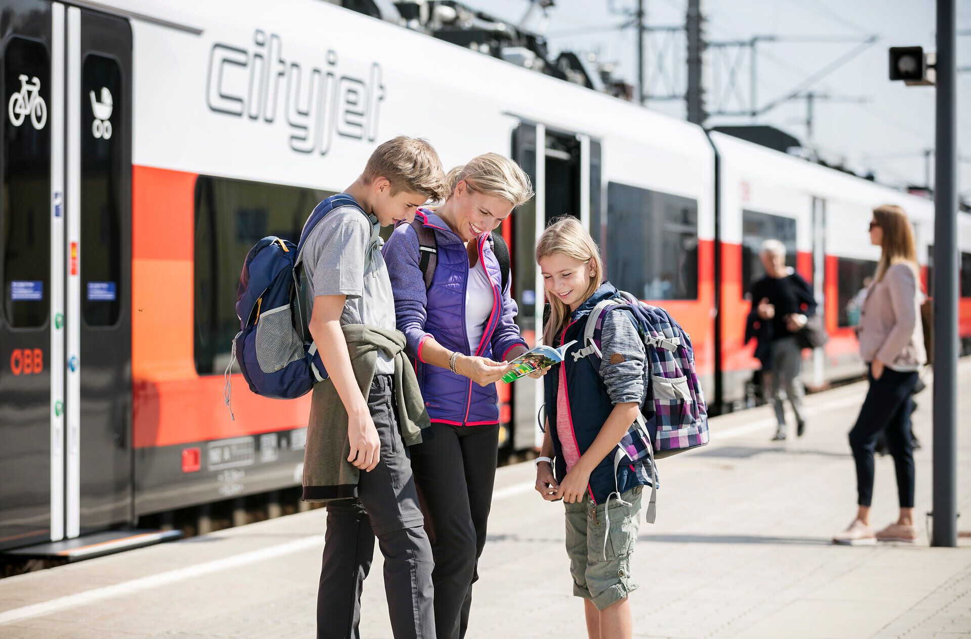 Eine fröhliche Familie steht am Bahnhof und studiert eine Karte, während sie auf ihren Zug wartet. Die Vorfreude auf das bevorstehende Abenteuer in den Bergen ist spürbar, umgeben von der lebhaften Atmosphäre des Bahnhofs.