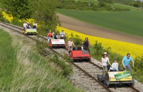 Menschen fahren auf Draisinen durch eine ländliche Landschaft mit gelben Rapsfeldern.