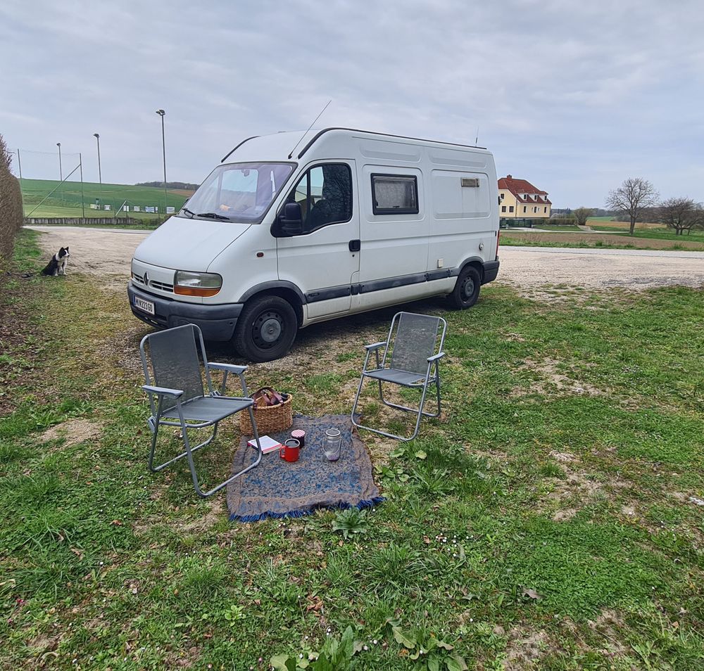 weißer Campingbus auf Schotteruntergrund mit grüner Wiese und Cmpingsessel im Vordergrund