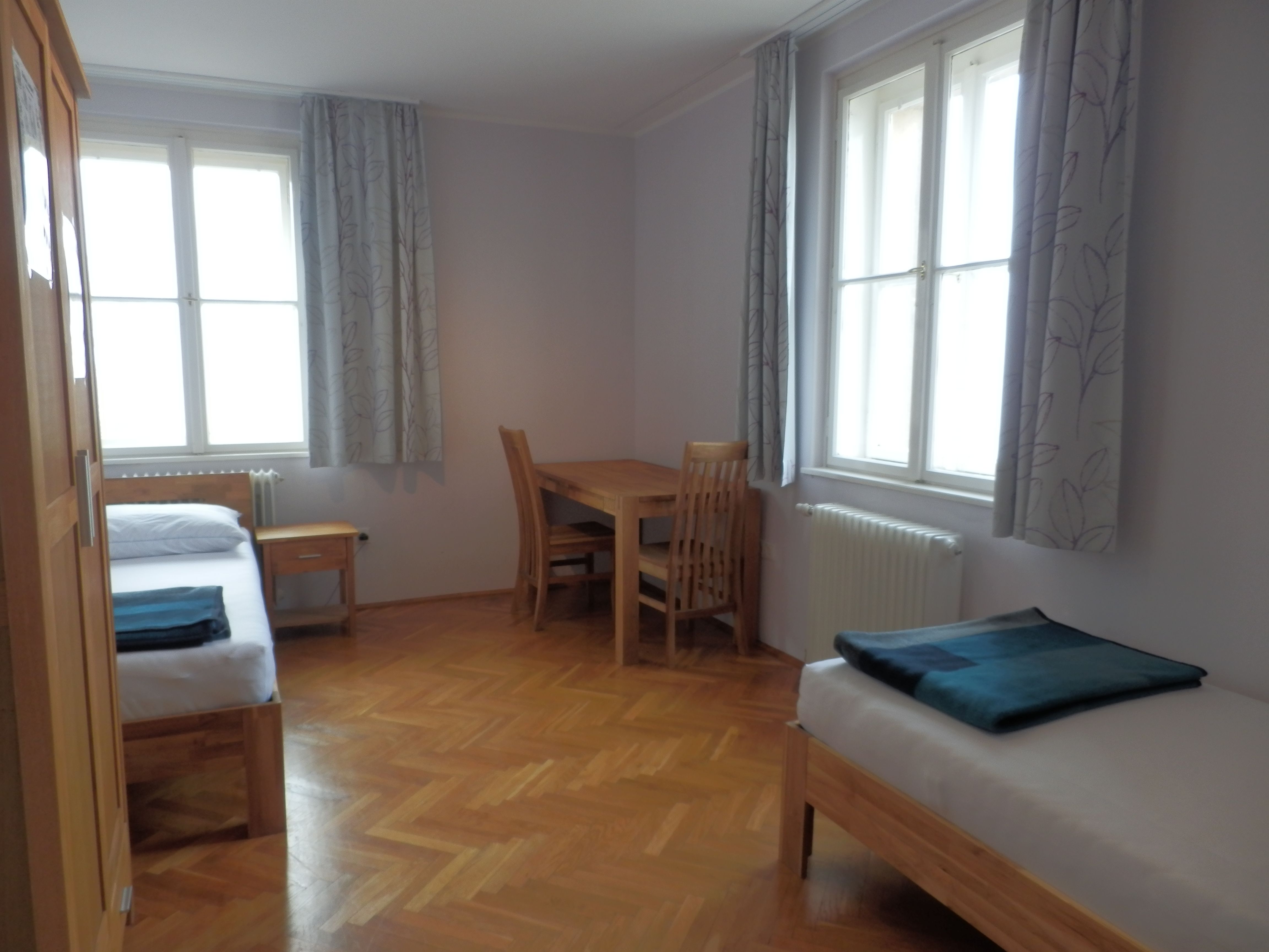 Ein einfach eingerichtetes Gästezimmer mit zwei Betten, einem Tisch und Stühlen, Holzboden und großen Fenstern mit Vorhängen.