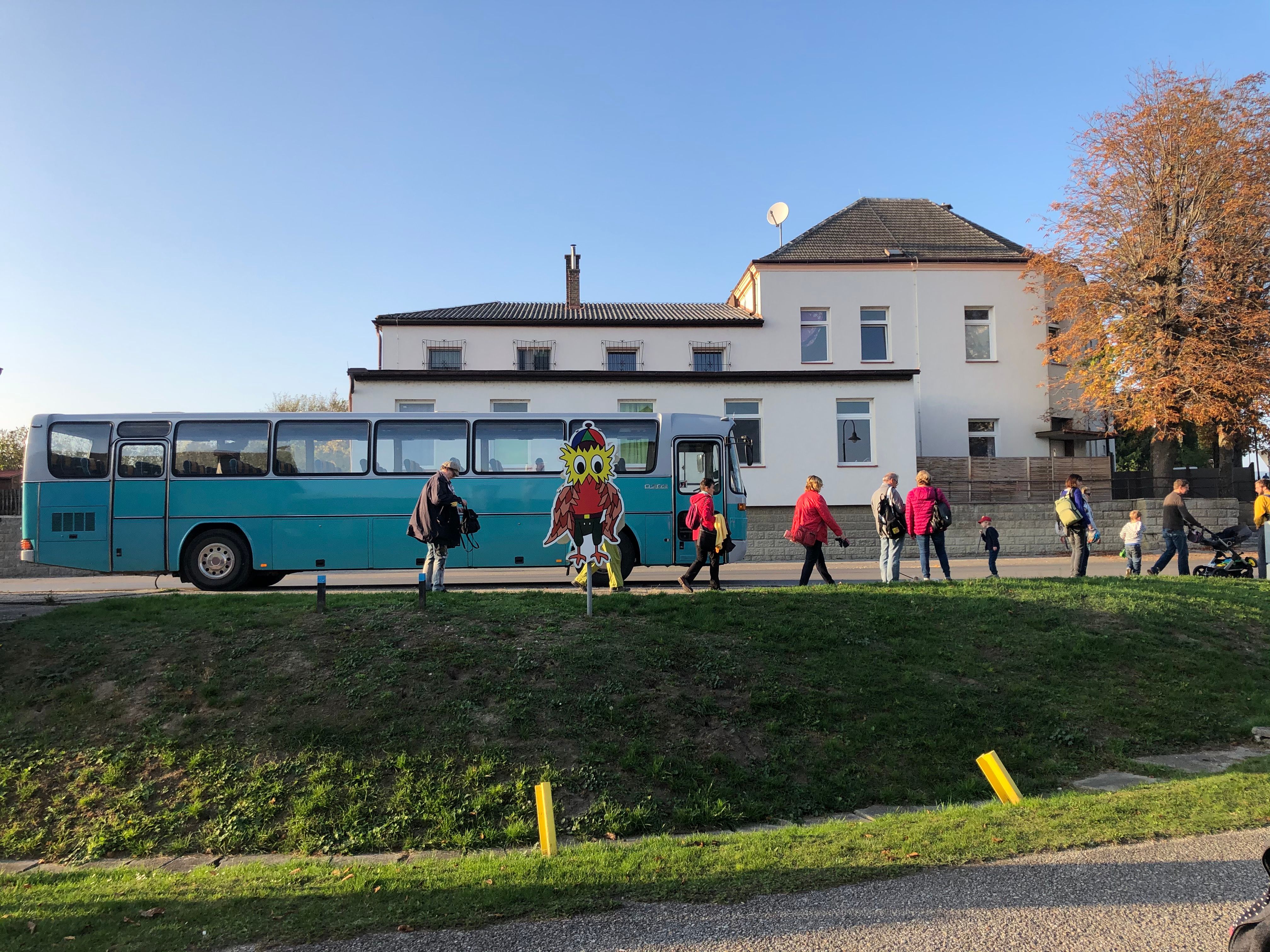 Ein blauer Bus steht vor einem Gebäude, Menschen gehen vorbei. Eine bunte Eulenfigur ist im Vordergrund zu sehen.