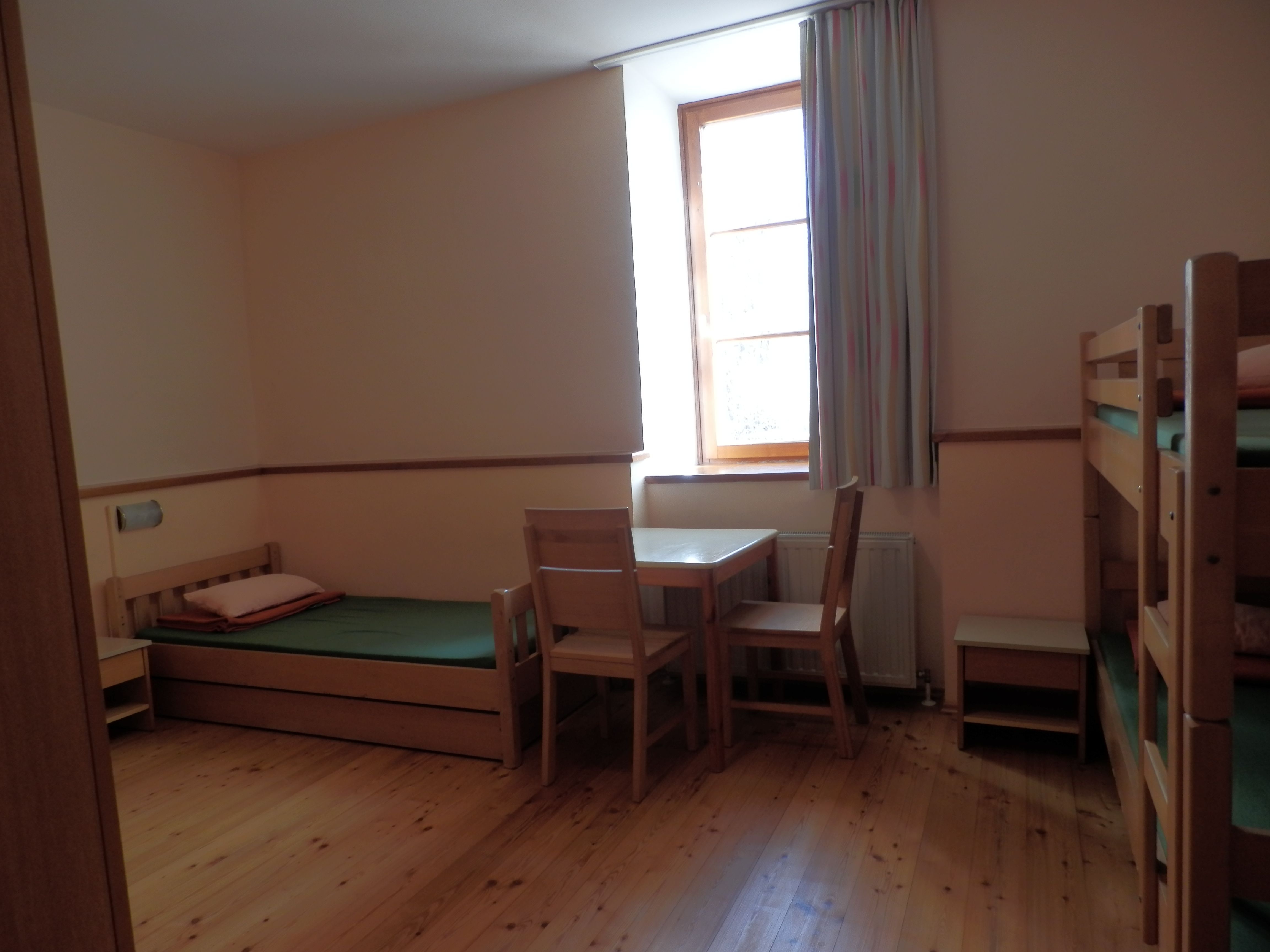 Ein einfach eingerichtetes Gästezimmer mit Holzboden, Einzelbett, Etagenbett, Tisch und Stühlen, sowie einem Fenster mit Vorhang.