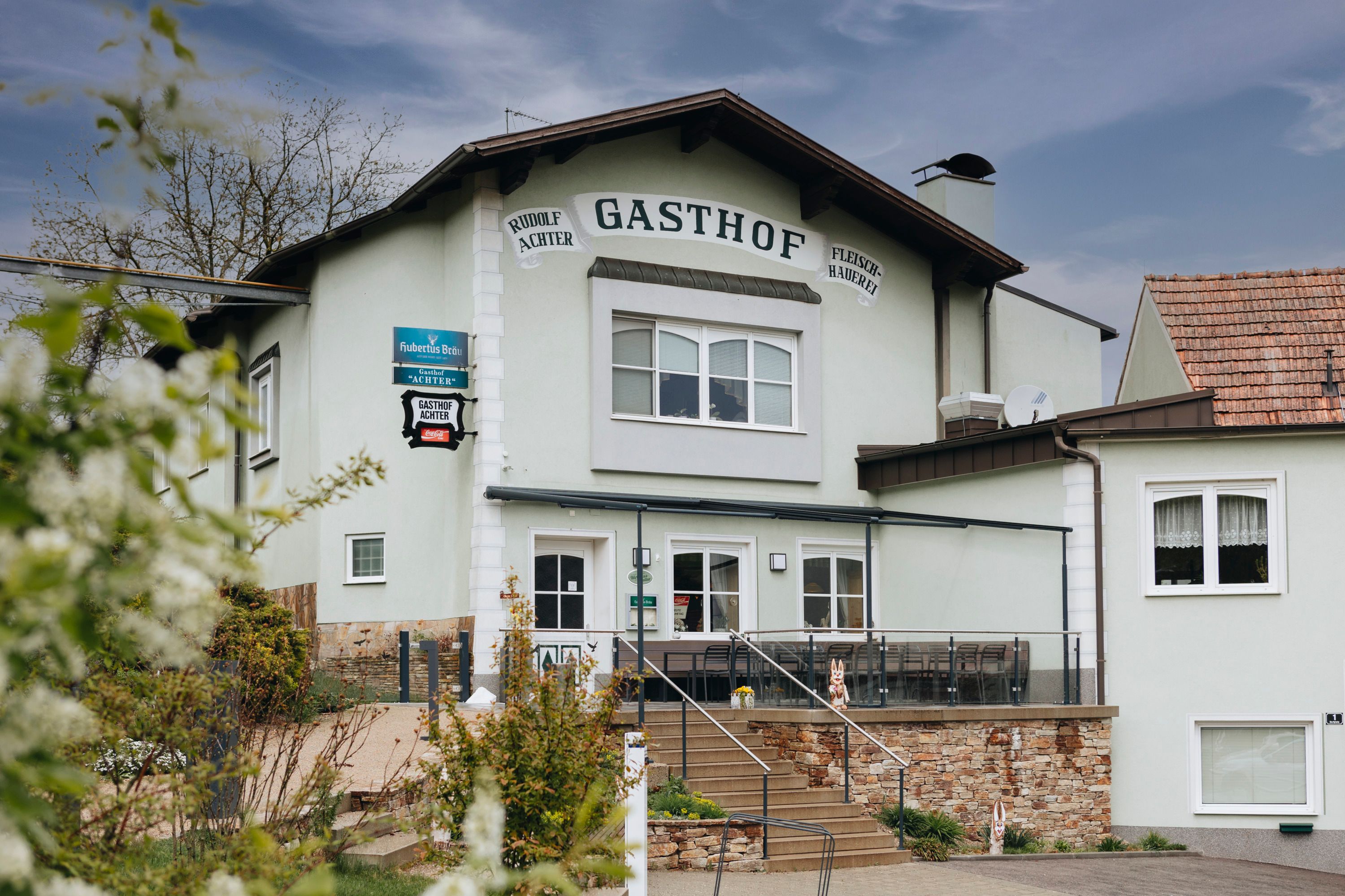 Außenansicht eines Gasthofs mit Terrasse und Treppe, umgeben von Pflanzen.