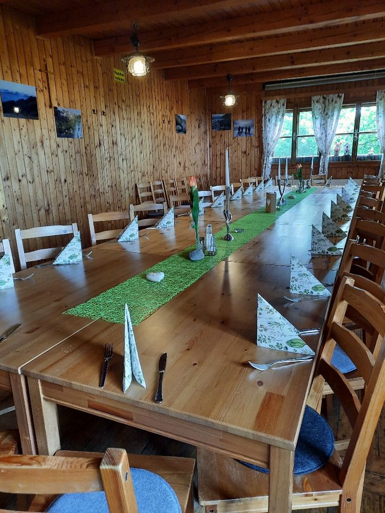 Ein rustikaler Raum mit Holzwänden und -decke. Ein langer Tisch ist mit grüner Tischdecke, Kerzen und gefalteten Servietten gedeckt.