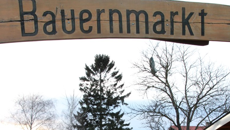 Entrance to a farmers' market with a wooden sign and a paved path surrounded by trees and buildings.