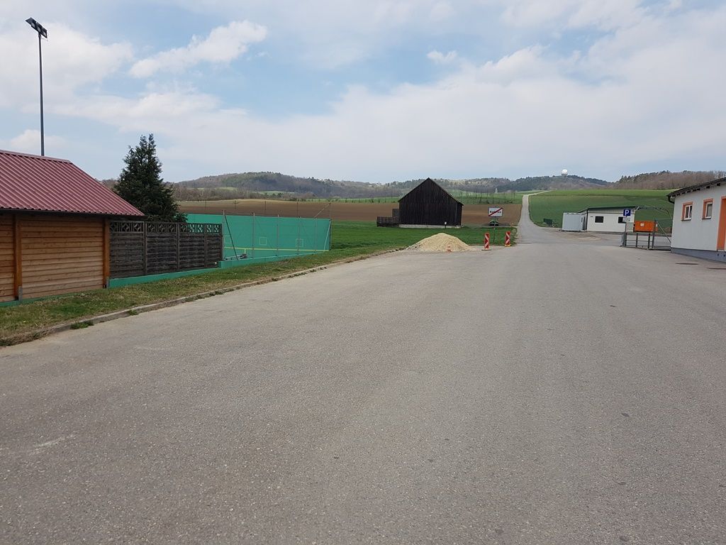 An empty parking lot with surrounding buildings and fields in the background.