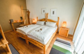 Ein gem&uuml;tliches Schlafzimmer mit Holzm&ouml;beln, einem Doppelbett, Nachttischen und einem Schreibtisch. Zwei Lampen und Bilder an der Wand erg&auml;nzen das Ambiente.