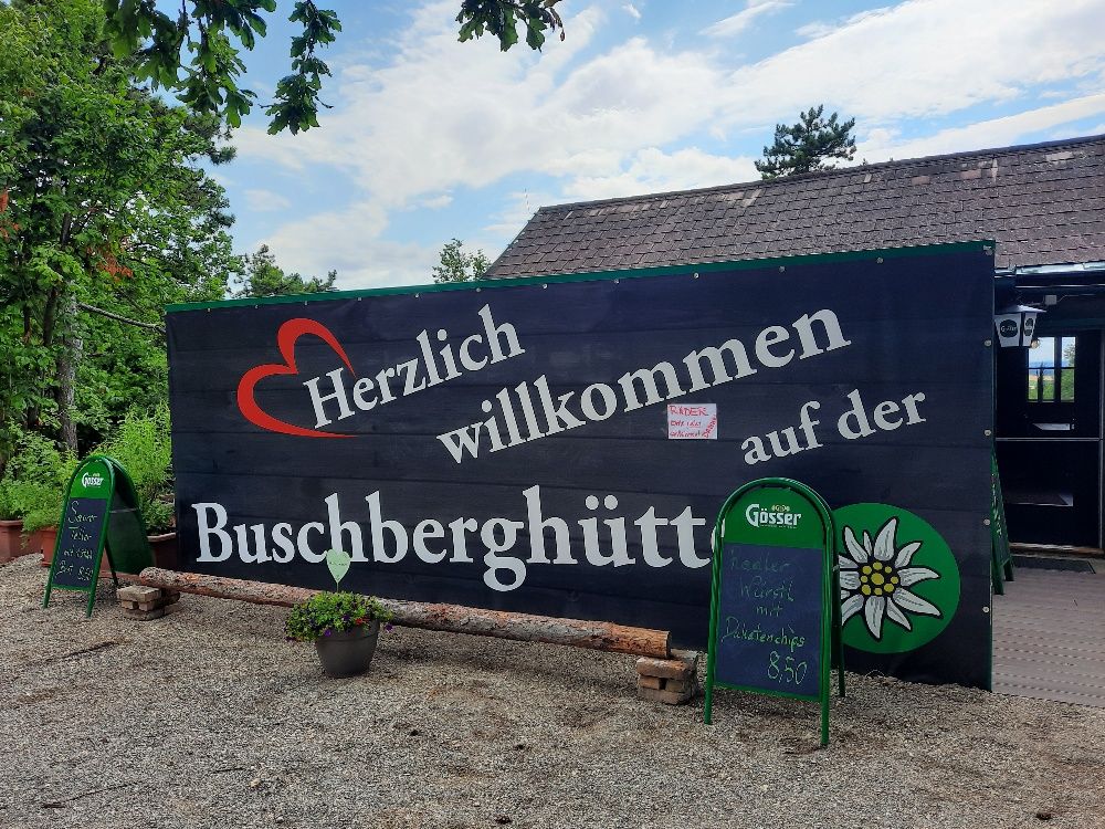 Willkommensschild der Buschberghütte mit grüner Umgebung und blauen Himmel.