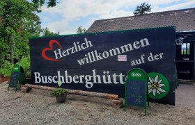 Willkommensschild der Buschberghütte mit grüner Umgebung und blauen Himmel.