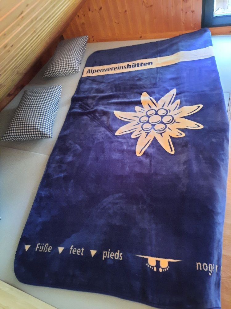 Ein Matratzenlager mit einer blauen Decke, die ein Edelweiß und den Schriftzug 'Alpenvereinshütten' zeigt, sowie zwei karierte Kissen.