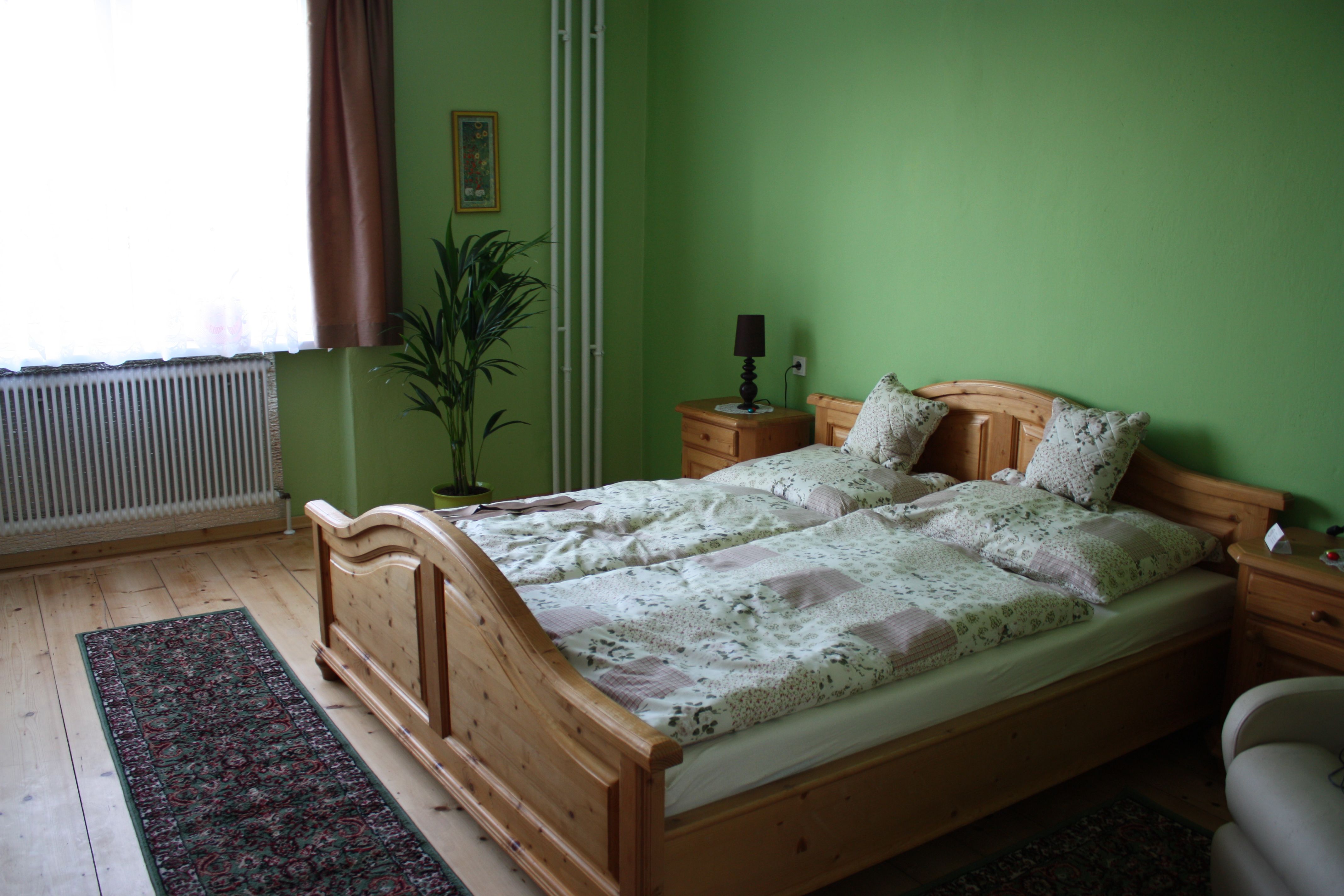 Ein gemütliches Gästezimmer mit grün gestrichenen Wänden, einem Holzbett, Nachttischen, einer Pflanze und einem Teppich auf Holzboden.