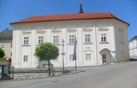 Historisches Gebäude mit rotem Dach und weißen Wänden, umgeben von Bäumen und einer Straße.
