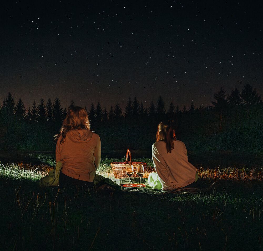 Unter dem funkelnden Sternenhimmel genießen zwei Freundinnen eine ruhige Nacht im Freien. Umgeben von der Stille der Natur und dem sanften Licht eines kleinen Picknicks, erleben sie einen unvergesslichen Moment der Verbundenheit und des Staunens.