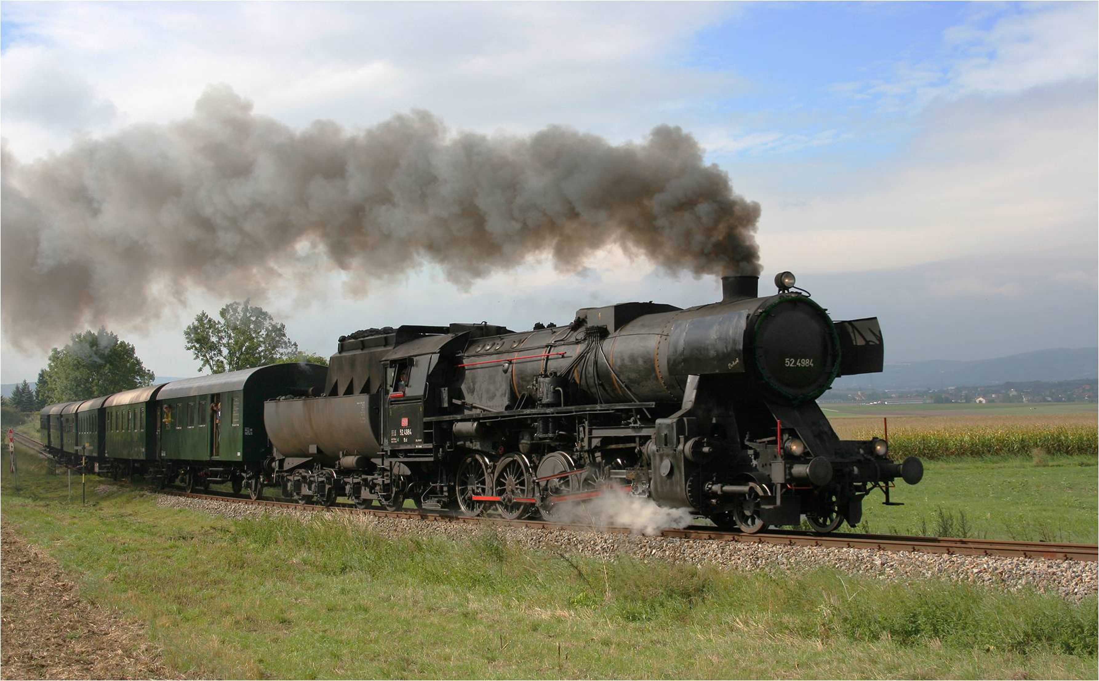 Eine dampfende, schwarze Dampflokomotive zieht grüne Waggons durch eine ländliche Landschaft.