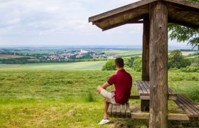 Ein Mann sitzt unter einem Holzunterstand und blickt auf eine weite Landschaft mit Feldern und einem Dorf in der Ferne.