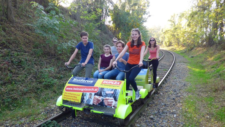 Gruppe von Menschen f&auml;hrt auf einer Draisine auf Bahngleisen durch eine bewaldete Landschaft.