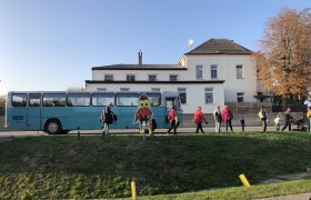 Ein blauer Bus steht vor einem Geb&auml;ude, Menschen gehen vorbei. Eine bunte Eulenfigur ist im Vordergrund zu sehen.