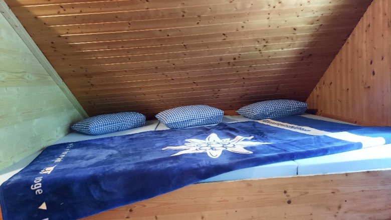 Ein Matratzenlager mit blauer Decke und karierten Kissen unter einer Holzdecke.