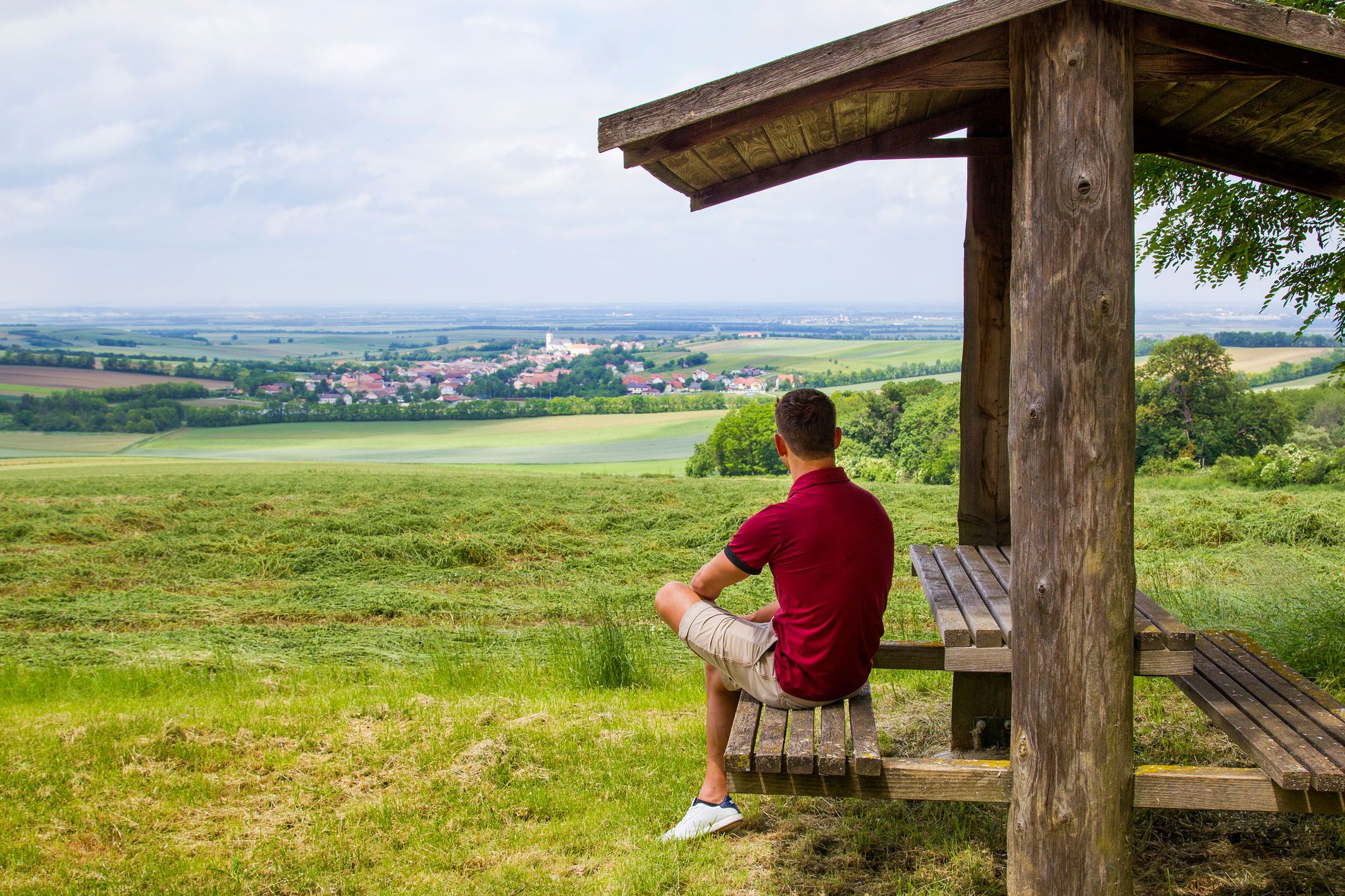 Ein Mann sitzt unter einem Holzunterstand und blickt auf eine weite Landschaft mit Feldern und einem Dorf in der Ferne.