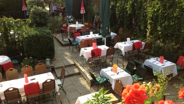 A cozy guest garden with covered tables, surrounded by green plants and red flowers.