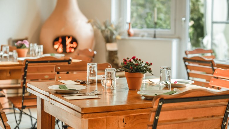 Ein gem&uuml;tlicher Fr&uuml;hst&uuml;cksraum mit Holztischen und -st&uuml;hlen, gedeckt mit Geschirr und Gl&auml;sern. Ein Kamin im Hintergrund sorgt f&uuml;r eine warme Atmosph&auml;re.