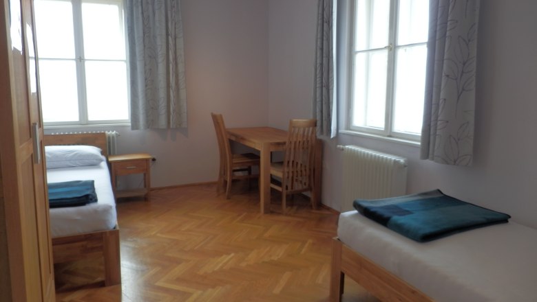 A simply furnished guest room with two beds, a table and chairs, wooden floor and large windows with curtains.