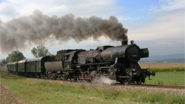 Eine dampfende, schwarze Dampflokomotive zieht grüne Waggons durch eine ländliche Landschaft.
