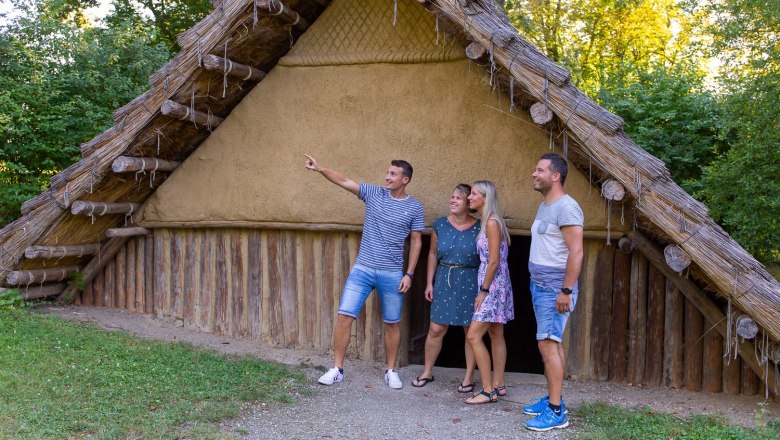 Four people stand in front of a reconstructed prehistoric house outdoors.