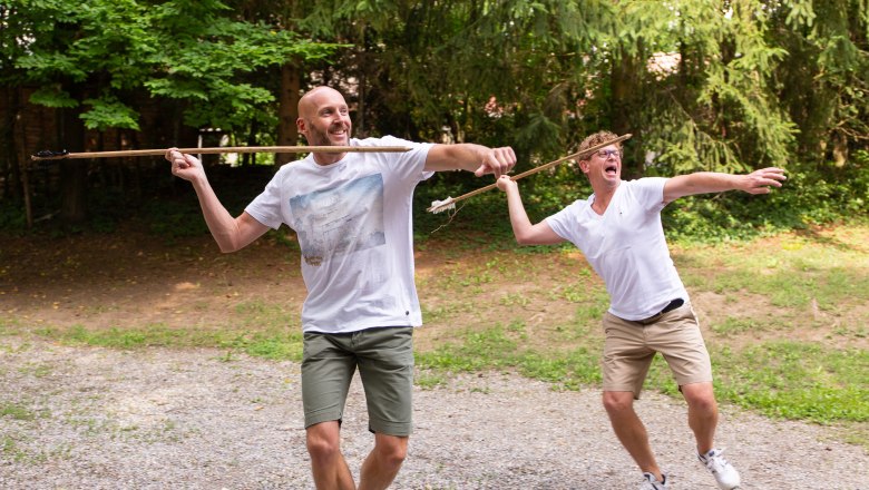 Two men throw spears outdoors, surrounded by trees.