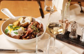 Ein Teller mit einem Gericht aus Fleisch, Kartoffeln und Spiegelei, serviert mit einem Glas Weißwein auf einem gedeckten Tisch.